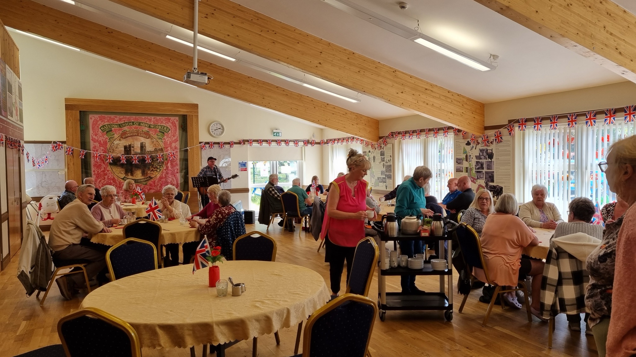 Older people sitting at round tables in a bright community hall, with a musician playing guitar and union flag bunting overhead