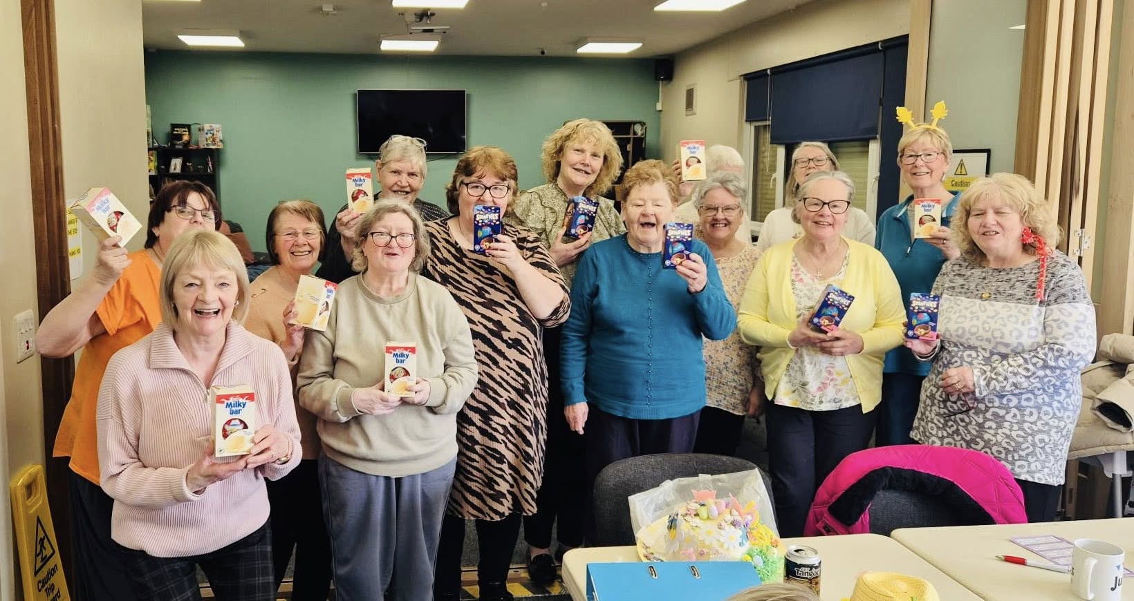 A group of smiling women holding up boxes of Easter eggs in a community room