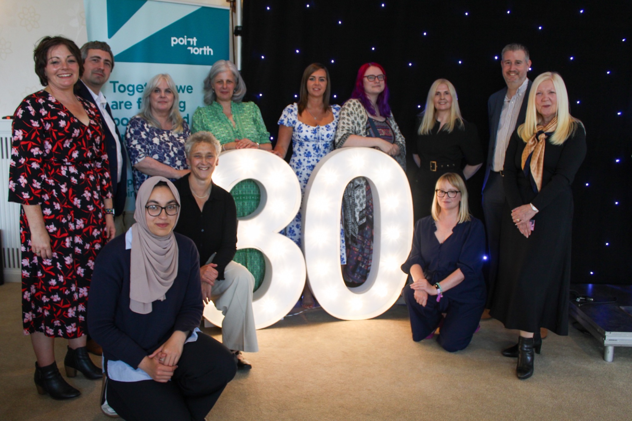 The Point North team gathered around illuminated 30 numerals beside a Point North banner